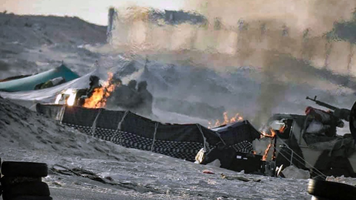 L'image semble représenter une scène de conflit ou de guerre, avec des flammes visibles et de la fumée s'élevant dans l'air. On peut distinguer des structures endommagées et une ambiance chaotique. Le sol est recouvert de débris, et des véhicules militaires ou des équipements peuvent être présents. L'environnement semble aride, suggérant un paysage désertique.