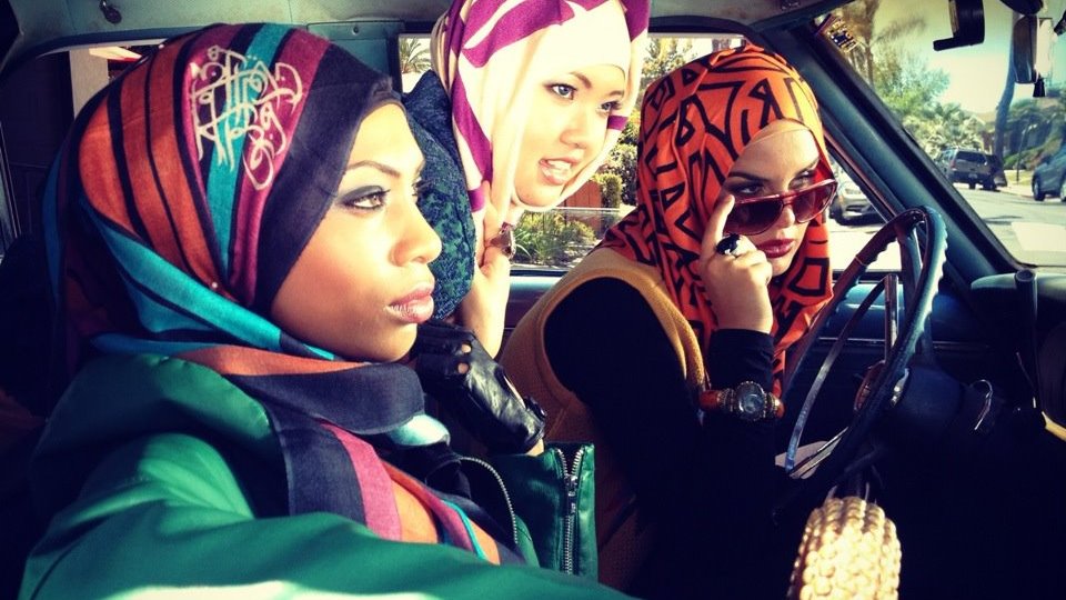 L'image montre trois femmes à l'intérieur d'une voiture. Elles portent des hijabs colorés avec des motifs variés. L'une d'elles est au volant, tandis que les deux autres semblent attentives à ce qui se passe à l'extérieur. L'ambiance est dynamique et moderne, mettant en valeur des styles vestimentaires tendance. Les couleurs vives des hijabs contrastent avec l'intérieur de la voiture, créant une atmosphère vibrante.