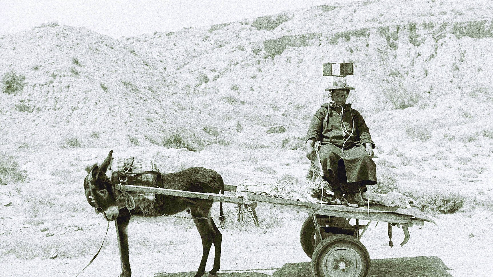 L'image montre une scène en noir et blanc d'un homme assis sur un chariot tiré par un âne. L'homme porte des vêtements traditionnels et semble se diriger quelque part, tandis que l'âne est attaché à l'avant du chariot. L'arrière-plan présente un paysage aride et désertique, avec des collines ou des montagnes à l'horizon. L'atmosphère de la photographie évoque un sentiment de simplicité et de vie rurale.