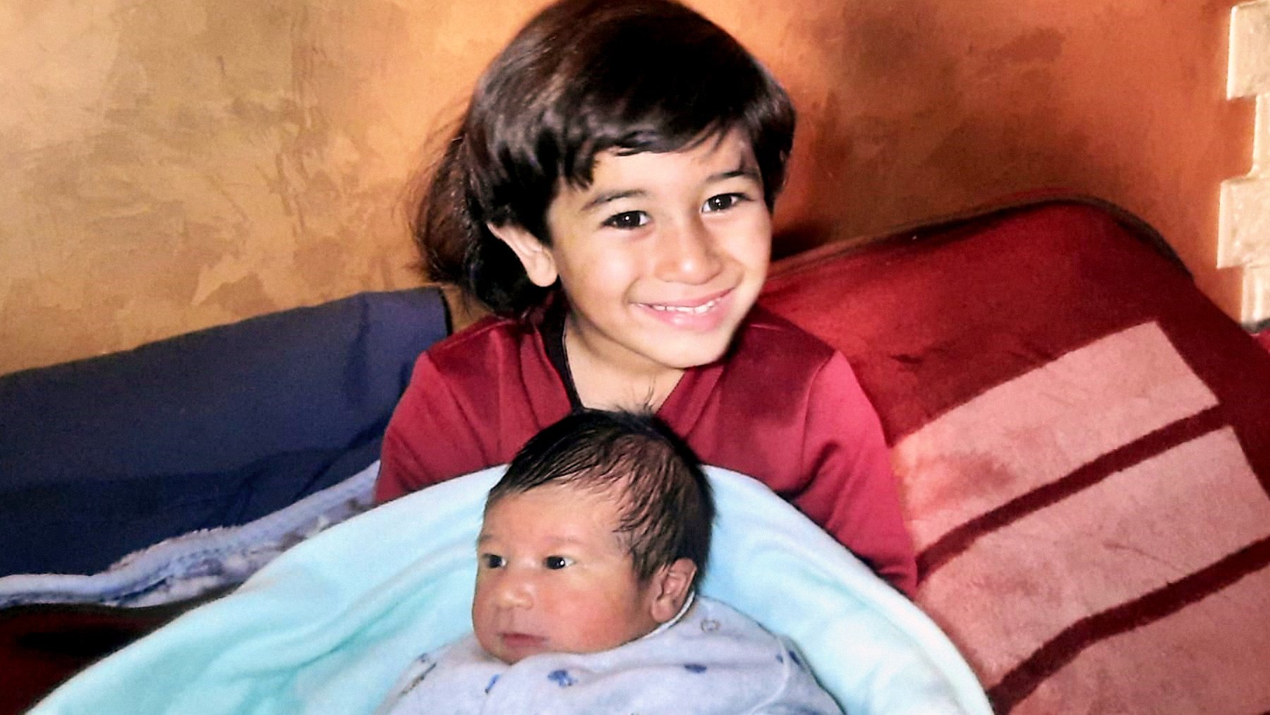 L'image montre un enfant souriant assis sur un canapé, tenant un bébé enveloppé dans une couverture bleue. L'enfant plus âgé semble heureux et fier, tandis que le bébé a une expression calme. Le décor est chaleureux avec des murs aux teintes terreuses.