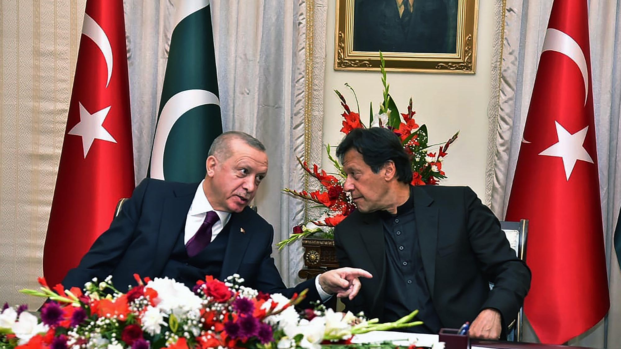 L'image montre deux hommes en conversation, assis à une table décorée de fleurs. En arrière-plan, on peut voir des drapeaux turc et pakistanais, ainsi qu'un portrait accroché au mur. Les hommes semblent échanger des idées de manière animée, et l'atmosphère semble conviviale.
