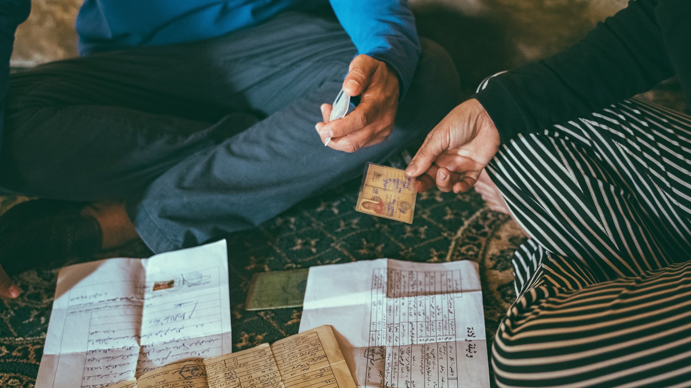 L'image montre deux personnes assises par terre, entourées de divers documents étalés devant elles. L'une des personnes tient une carte d'identité ou un document similaire, tandis que l'autre semble utiliser un marqueur pour écrire ou annoter quelque chose. Les documents sur le sol sont variés, comprenant des papiers manuscrits et des feuilles à imprimer. L'environnement semble intime et informel, suggérant une discussion importante ou une transaction en cours.