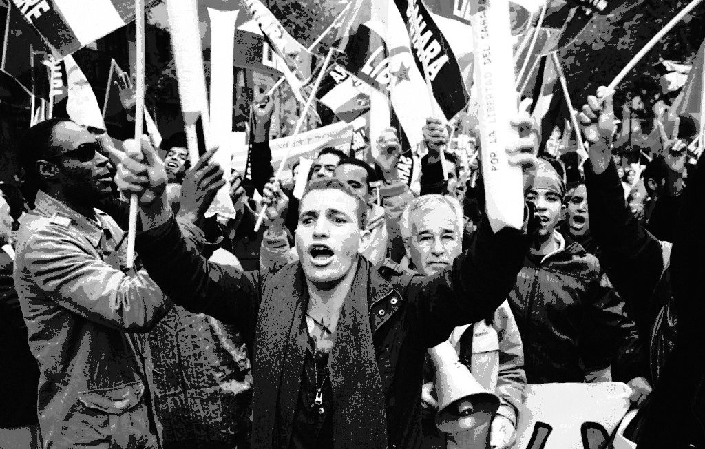 L'image montre une foule en pleine manifestation. On peut voir des personnes brandissant des drapeaux et des pancartes, exprimant leurs opinions avec passion. Au premier plan, un jeune homme s'écrie, tandis que d'autres personnes autour de lui semblent également mobilisées et engagées. L'ambiance générale dégage une forte énergie et un sentiment de solidarité parmi les manifestants. Les visages reflètent une gamme d'émotions, allant de la détermination à l'enthousiasme.