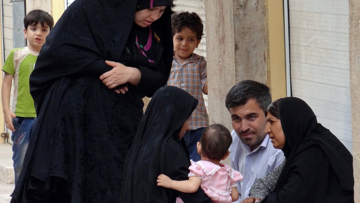L'image montre un groupe de personnes dans une rue. Deux femmes, vêtues de vêtements noirs, sont assises sur le sol et discutent. L'une d'elles tient un bébé, qui porte une légère robe rose. À proximité, un homme regarde les femmes et semble participer à la conversation. Un jeune garçon, en arrière-plan, observe la scène. L'environnement apparaît urbain, avec des bâtiments en briques visibles. L'atmosphère semble conviviale et familiale.