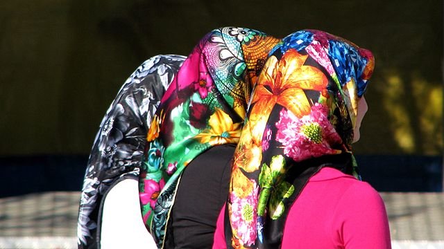 L'image montre le dos de trois femmes portant des foulards colorés. Chaque foulard est orné de motifs fleuris et vibrants, offrant un contraste frappant avec leurs vêtements. Les cheveux des femmes sont couverts, laissant entrevoir seulement leurs silhouettes. L'ambiance semble vivante et pleine de couleurs, soulignant la richesse culturelle des accessoires.