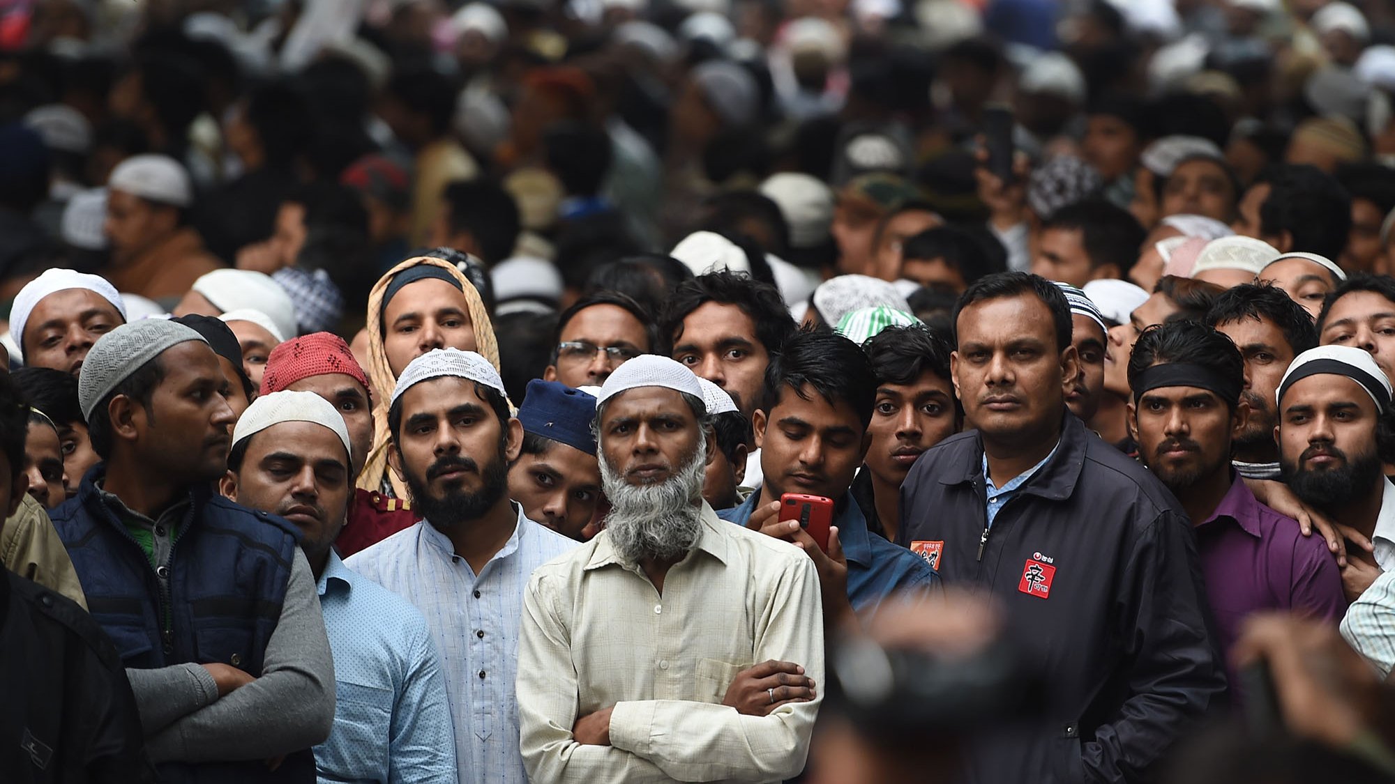 L'image montre une grande foule d'hommes, principalement vêtus de vêtements traditionnels, avec plusieurs d'entre eux portant des couvre-chefs. Les expressions des gens varient, certains semblent attentifs, tandis que d'autres regardent vers l'avant. L'ambiance dégage un sentiment de concentration et de rassemblement, possiblement pour un événement ou une cérémonie.