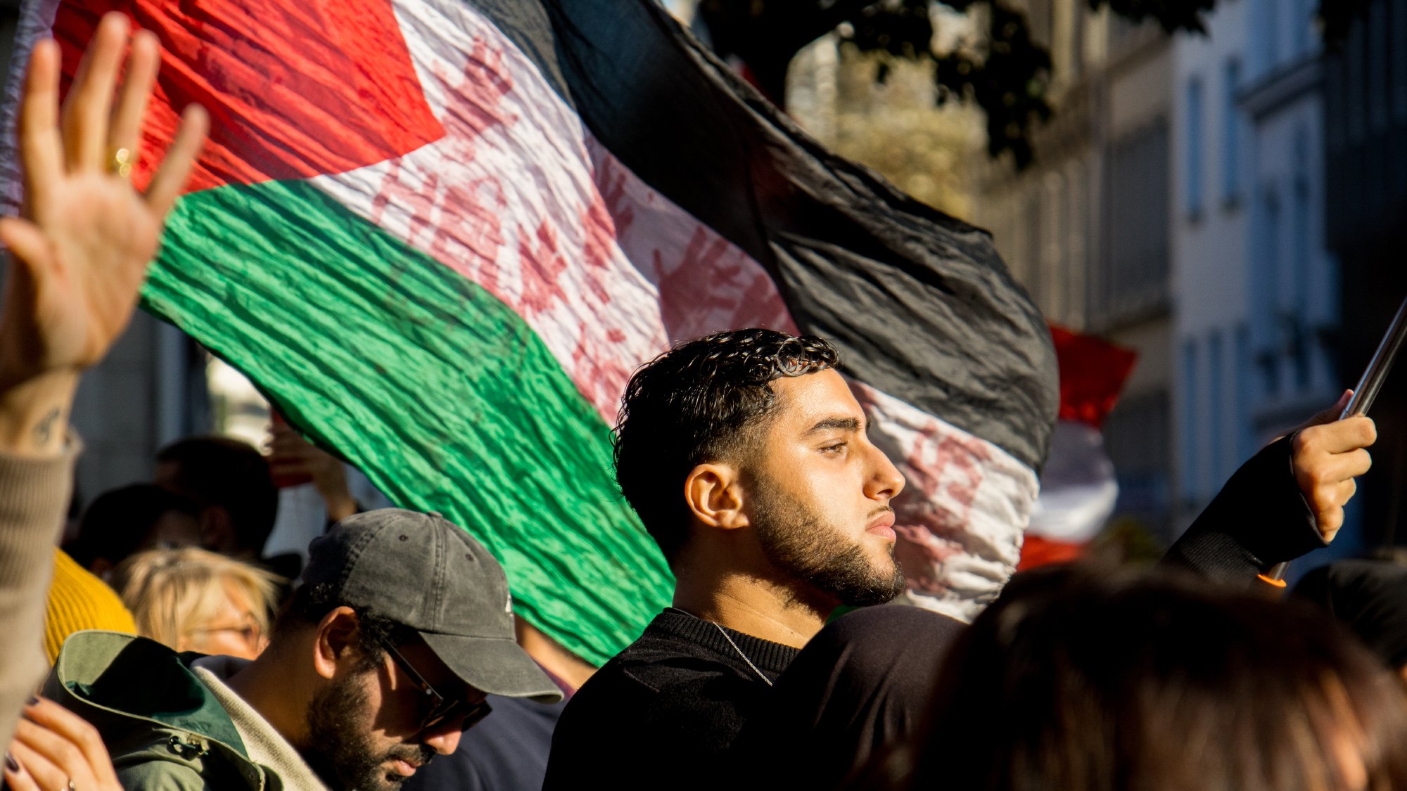 L'image montre une foule lors d'une manifestation. On peut voir une personne au premier plan tenant un grand drapeau aux couleurs du pays. Il y a également d'autres manifestants en arrière-plan, dont certains portent des lunettes de soleil et d'autres ont des expressions engagées. La lumière du soleil semble créer une ambiance dynamique et engagée, accentuant l'émotion de l'événement.