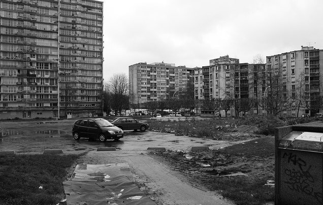 L'image montre un paysage urbain qui semble désolé. On peut voir plusieurs immeubles de grande hauteur, typiques de l'architecture des banlieues. Le sol est quelques peu inégal, avec des flaques d'eau et des débris éparpillés, ce qui donne une impression de négligence. Deux voitures sont garées sur un chemin de terre. L'environnement est sombre, accentué par le ciel nuageux, ce qui ajoute à l'atmosphère générale de tristesse et de délabrement.