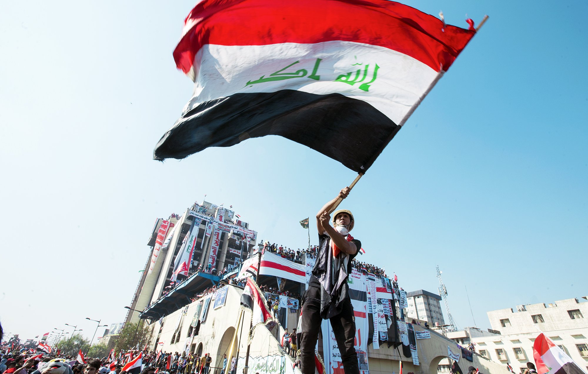 L'image montre une personne agitant un drapeau irakien. Le drapeau, aux couleurs rouge, blanc, noir et vert, est bien visible. La scène se déroule probablement lors d'une manifestation ou d'un événement public, avec des bâtiments en arrière-plan décorés de drapeaux. L'atmosphère semble dynamique, reflétant un sentiment de patriotisme et de rassemblement.