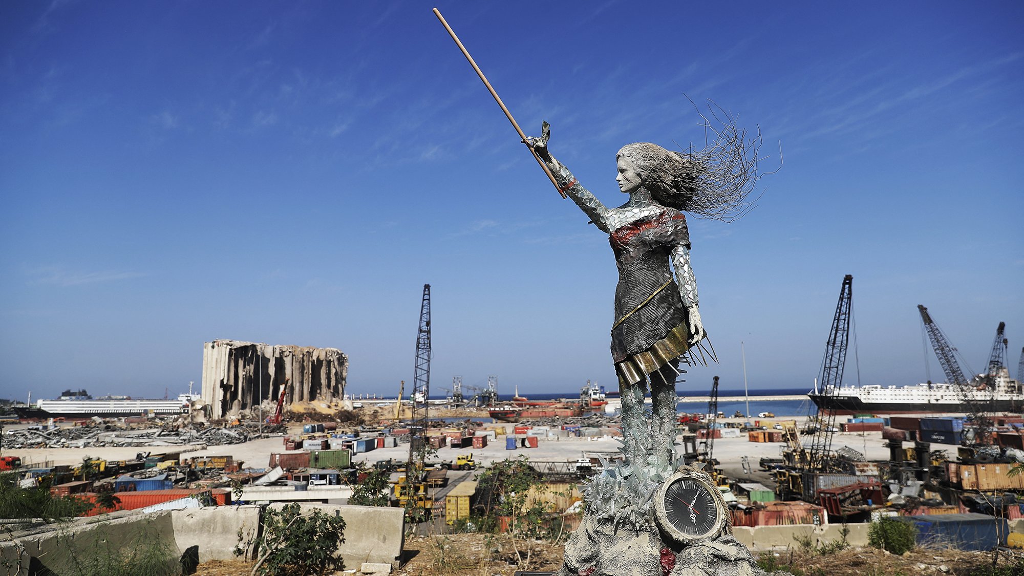 L'image présente une sculpture en métal d'une femme levant un bâton, symbolisant probablement la force et la résilience. Elle se tient sur une plateforme, surplombant un paysage industriel ou portuaire, avec des conteneurs, des grues et des bâtiments en arrière-plan. On peut également apercevoir des vestiges d'un bâtiment détruit, suggérant une scène de reconstruction ou de renouveau après une catastrophe. Le ciel est dégagé et ensoleillé, contrastant avec l'environnement chaotique du port.