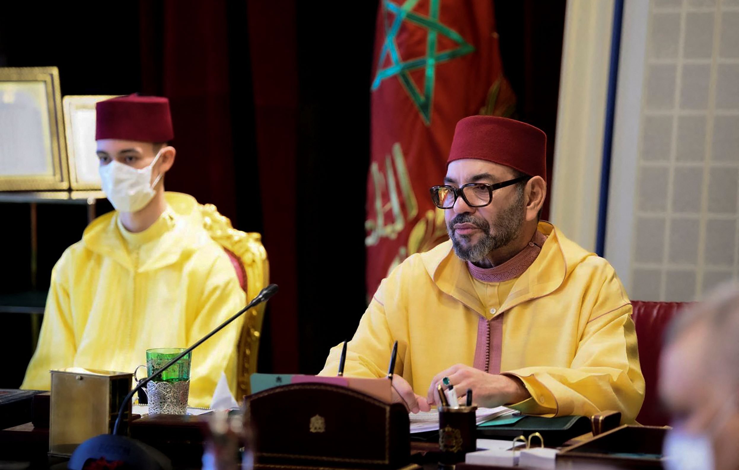L'image montre une réunion se déroulant dans un cadre officiel. À droite, un homme vêtu d'une tenue traditionnelle marocaine de couleur jaune, portant un fez rouge, est attentif et semble concentré sur les discussions. À gauche, un jeune homme portant également un jaune et un masque facial regarde devant lui. En arrière-plan, on peut apercevoir des éléments de décoration et des drapeaux, évoquant le contexte marocain de la scène. L'ambiance générale semble sérieuse et solennelle.