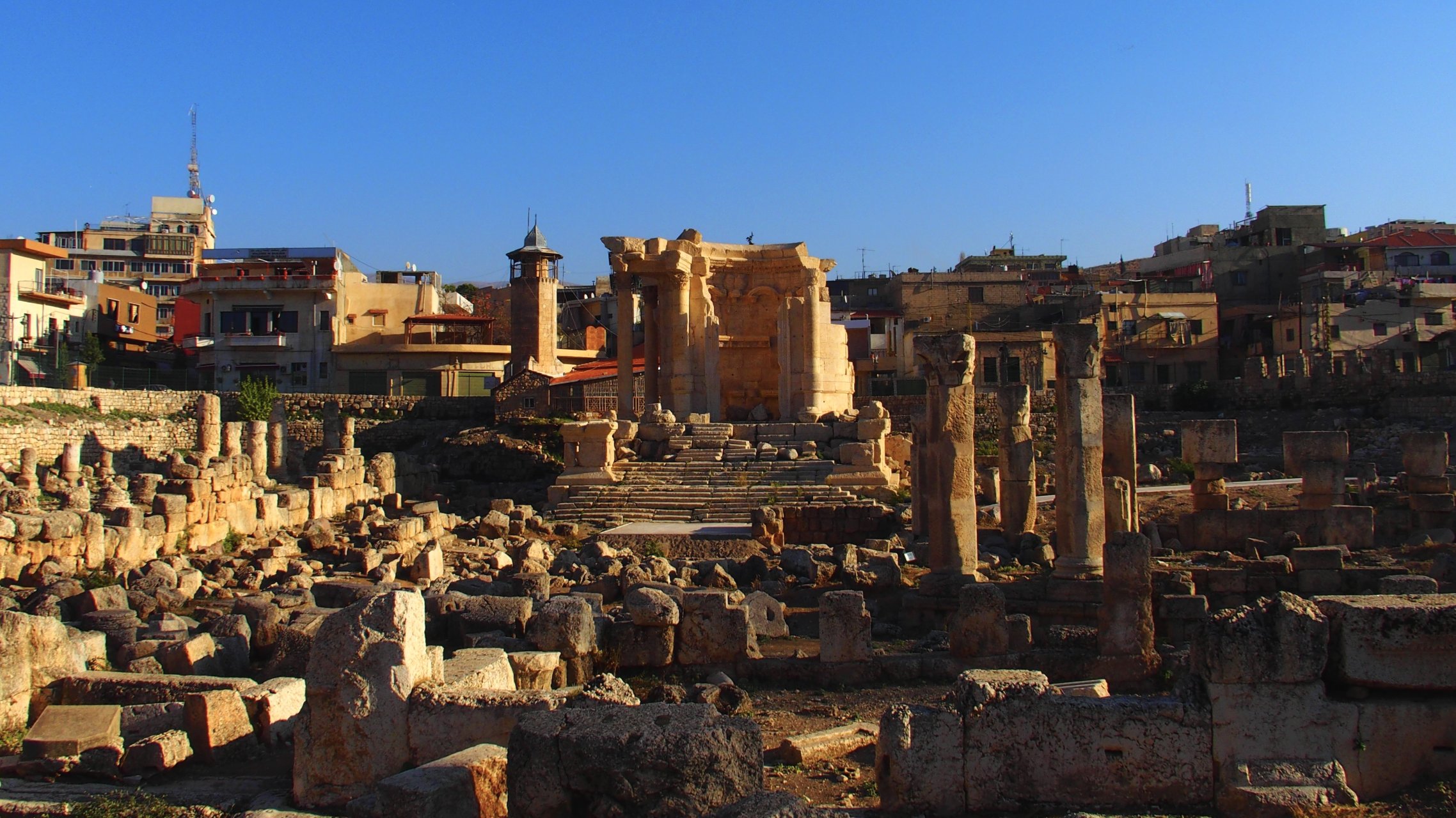 L'image montre un site archéologique avec des ruines anciennes, probablement d'un temple, entourées de colonnes et de blocs de pierre. En arrière-plan, on voit des bâtiments modernes et des structures urbaines s'élevant sous un ciel bleu. La lumière du soleil éclaire les ruines, créant un contraste entre l'ancien et le moderne. Ce lieu évoque une riche histoire et une importance culturelle.