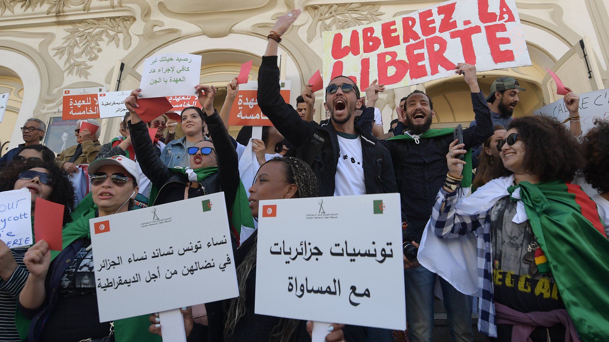 L'image montre un groupe de personnes rassemblées lors d'une manifestation. Les manifestants portent des pancartes et des drapeaux, exprimant des slogans tels que "Libérez la liberté". Les participants semblent engagés et passionnés, brandissant des affiches avec des messages en arabe et en français, appelant à la justice et à l'égalité. L'ambiance est énergique et déterminée, témoignant d'une dynamique de revendication sociale ou politique. L'arrière-plan présente une architecture caractéristique, ajoutant au contexte de l'événement.