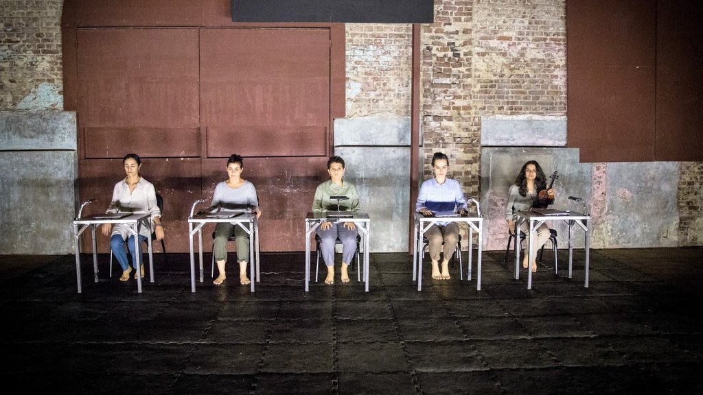 L'image montre un groupe de cinq personnes assises sur des chaises, disposées en ligne. Elles portent des vêtements simples et sont pieds nus, ce qui donne une impression de simplicité et d'intimité. L'environnement est industriel, avec des murs en brique apparente et une lumière tamisée qui crée une ambiance à la fois contemplative et moderne. Les personnes semblent concentrées, chacune avec un objet devant elle, suggérant un moment de performance ou de réflexion partagée.
