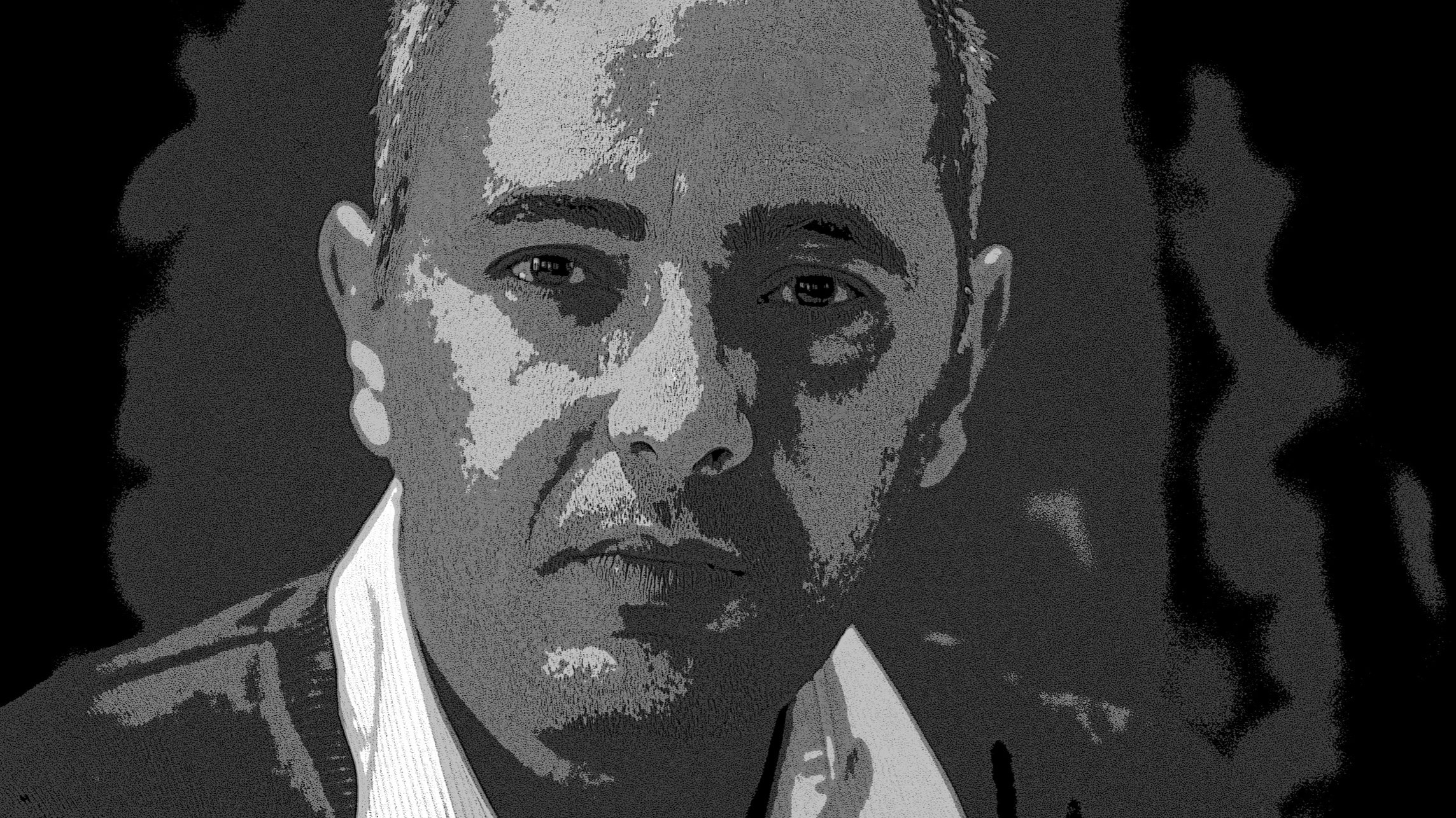 L'image montre un homme avec une expression réfléchie. Il a les cheveux courts et porte un pull. Ses mains sont croisées devant lui. Le fond est sombre, ce qui met en avant son visage. L'éclairage accentue ses traits et donne une atmosphère sérieuse à la photo.