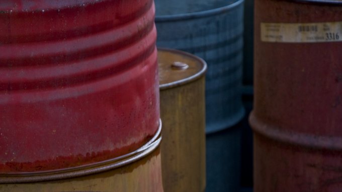 L'image montre plusieurs fûts en métal empilés les uns sur les autres. Chaque fût a une couleur différente : rouge, vert, noir et jaune. La surface des fûts semble briller, probablement à cause d'un liquide qui s'y trouve, reflétant la lumière. L'ensemble de la composition donne une impression de dépôt industriel, avec une texture rustique et des nuances de couleurs intéressantes.