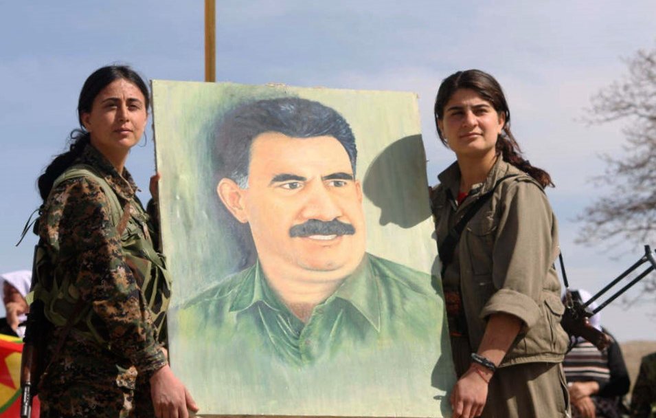 L'image montre deux femmes en tenue militaire tenant un portrait d'un homme avec une moustache. Elles semblent participer à un événement ou une manifestation, entourées d'une atmosphère qui indique une forme de dévotion ou de commémoration. Le fond est naturel, avec un ciel partiellement nuageux. Les expressions des femmes suggèrent détermination et fierté.