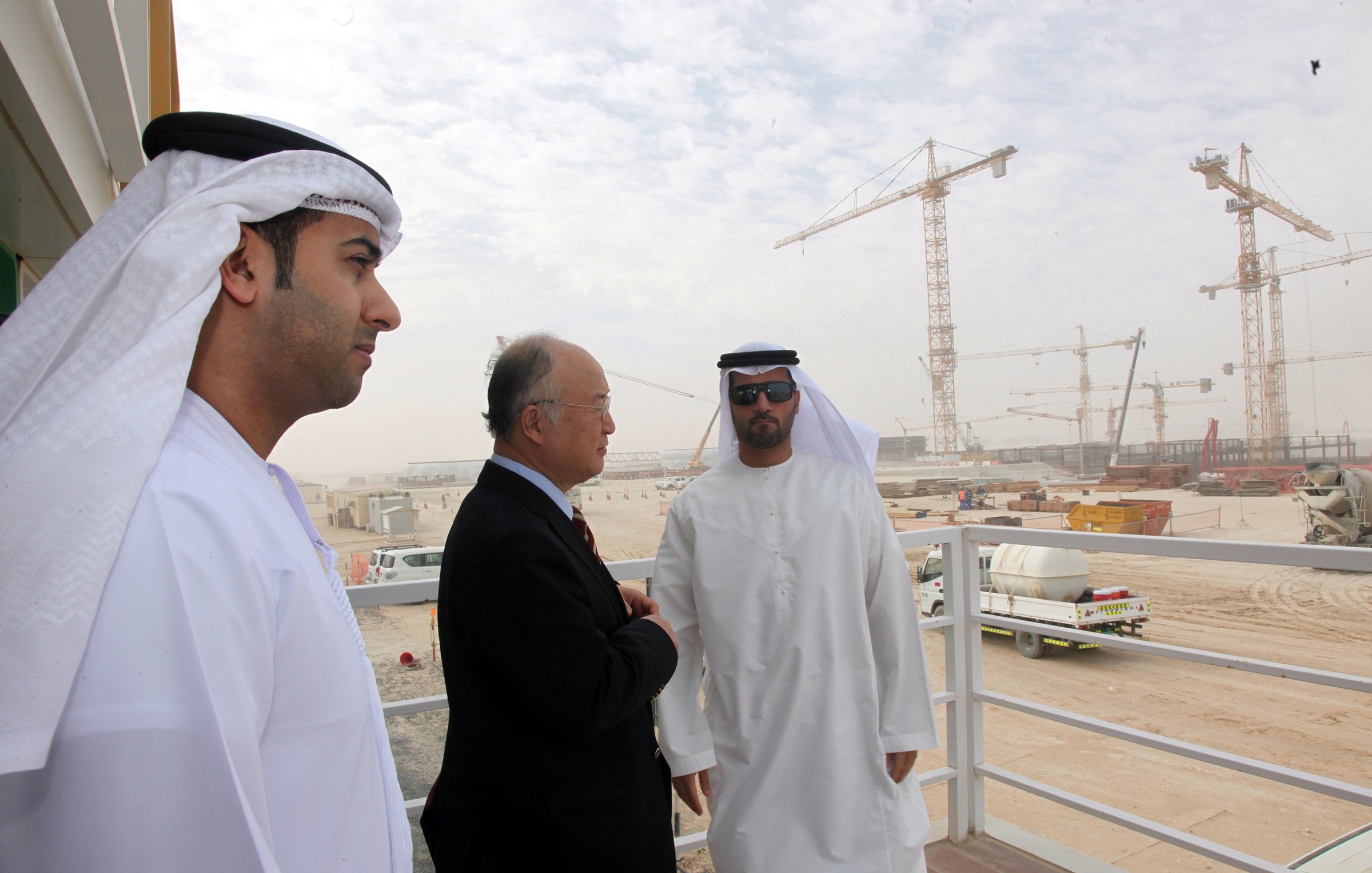 L'image montre trois hommes, apparemment en discussion, sur un balcon ou une plateforme. Ils sont vêtus de vêtements traditionnels des Émirats, comprenant des kandouras et des ghutras. En arrière-plan, on peut voir un site de construction avec des grues et des véhicules, suggérant un projet de développement en cours. L'environnement est poussiéreux et légèrement brumeux, typique des zones de construction dans des climats chauds. Les hommes semblent sérieux et engagés dans leur conversation.
