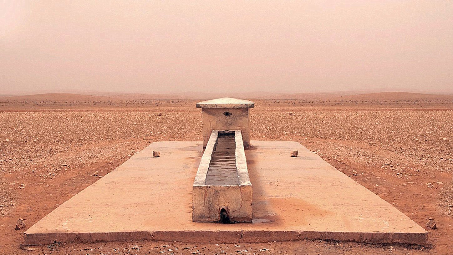 L'image montre une structure isolée située dans un paysage désertique. Au centre, il y a un long bassin en pierre, entouré par une surface plane et aride. Le sol est de couleur beige et semble sec et craquelé, tandis que l'atmosphère est légèrement brumeuse, ce qui donne une impression de chaleur et d'immensité désertique. Au loin, on aperçoit des collines ou des dunes qui se fondent avec le ciel dans une teinte douce et uniforme. L'endroit dégage une sensation de tranquillité et de solitude.