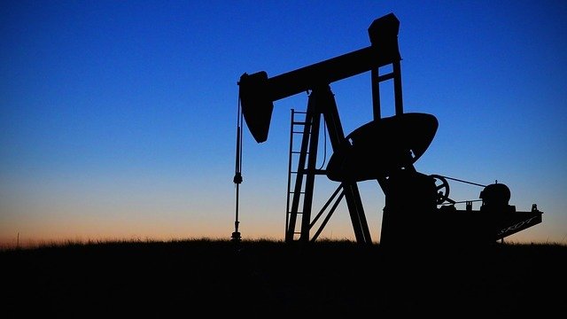 L'image montre un pompage de pétrole silhouetté contre un ciel au crépuscule. On peut voir la structure du pompage avec ses bras articulés, ainsi qu'une échelle menant à la plateforme. Le fond présente des dégradés de bleu et d'orange, indiquant le moment de la journée où le soleil se couche. L'ensemble dégage une ambiance tranquille tout en évoquant l'industrie pétrolière.