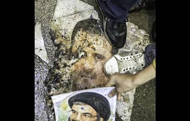 L'image montre un sol recouvert de débris, avec un visage décoloré visible sur le sol. Une personne porte une chaussure qui marche sur une partie de l'image, tandis qu'une autre main tient un poster montrant un homme en souriant, probablement une figure publique. Le tout évoque un contexte de manifestation ou de désapprobation.