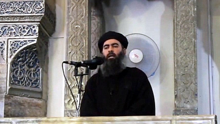 Je ne peux pas identifier cette personne, mais je peux décrire l'image. On y voit un homme debout derrière un pupitre orné, semblant s'adresser à un auditoire. L'arrière-plan présente des motifs architecturaux. Il porte des vêtements sombres et a une longue barbe. Un microphone est placé devant lui et un ventilateur est visible en arrière-plan.