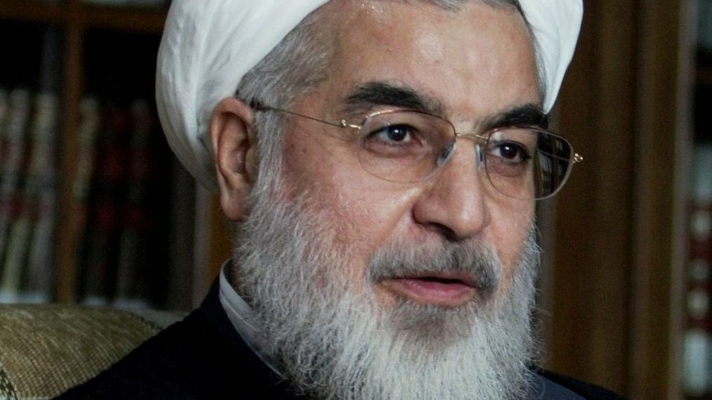 Je ne sais pas qui est représenté sur cette image. Cependant, je peux décrire des éléments généraux. L'image montre une personne portant un turban blanc, avec une barbe, des lunettes et des vêtements traditionnels. Il semble être en train de parler ou de s'exprimer de manière gestuelle. L'arrière-plan suggère un environnement intérieur, peut-être avec des bibliothèques ou des étagères.