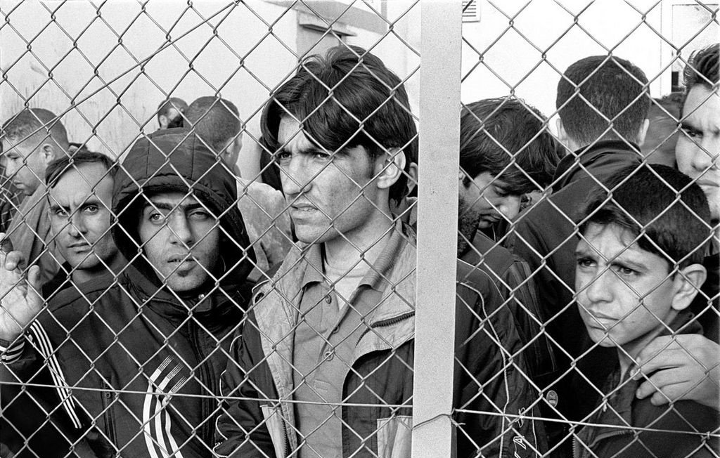 L'image montre un groupe d'hommes, visiblement en attente, qui se trouvent derrière une clôture en fil de fer. Leur expression faciale révèle une certaine anxiété et tension. Ils semblent chercher à voir ou à communiquer avec le monde extérieur. Le cadre en noir et blanc accentue l'intensité de leurs émotions et la gravité de la situation.