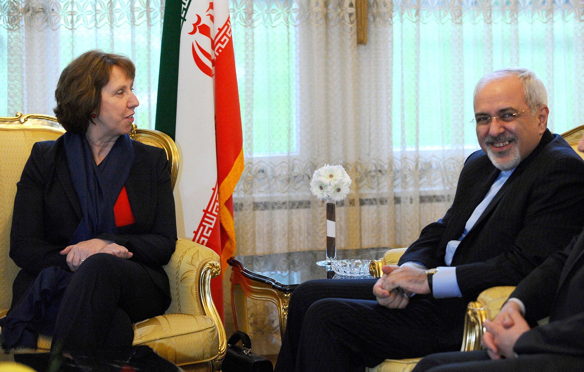 L'image montre une rencontre entre deux personnes assises sur des fauteuils dans un décor formel. À gauche, une femme avec des cheveux bruns, portant un blazer noir et un haut rouge, semble engagée dans la conversation. À droite, un homme souriant, vêtu d'un costume sombre, répond à la femme. En arrière-plan, on peut voir un drapeau iranien, ce qui suggère que la rencontre a lieu en Iran. La lumière naturelle entre par les fenêtres, créant une atmosphère accueillante.