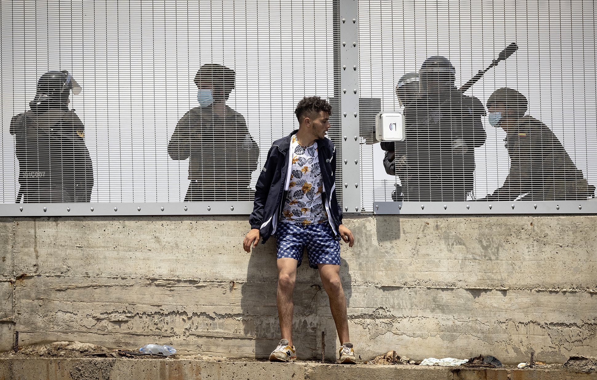 L'image montre un jeune homme debout contre un mur, habillé d'un haut sombre et d'un short à motifs. Il semble être en train de faire face à une situation tendue. Derrière lui, à travers une clôture en métal, on aperçoit des silhouettes de personnes en uniforme, apparemment des agents de la sécurité, qui semblent se préparer à intervenir. L'atmosphère est intense et suggère un contexte de conflit ou de protestation. Les ombres des agents renforcent le sentiment de pression ou de confrontation.