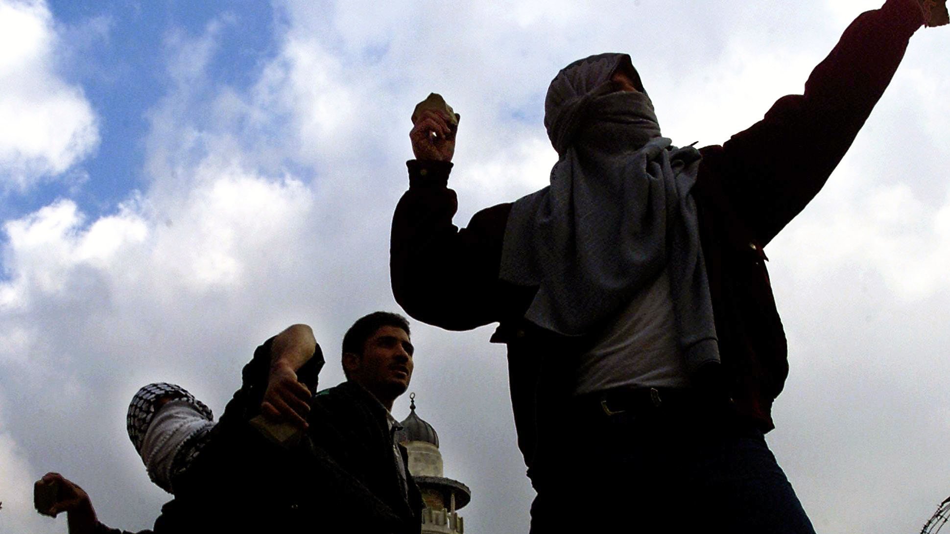 L'image montre plusieurs personnes en train de lancer des projectiles, probablement dans un contexte de confrontation ou de manifestation. Ils portent des vêtements sombres et ont le visage partiellement couvert, ce qui suggère qu'ils cherchent à dissimuler leur identité. En arrière-plan, on aperçoit une tour, probablement une mosquée, et des barbelés, renforçant l'idée d'un environnement tendu ou conflictuel. Le ciel est nuageux, ce qui contribue à l'atmosphère dramatique de la scène.