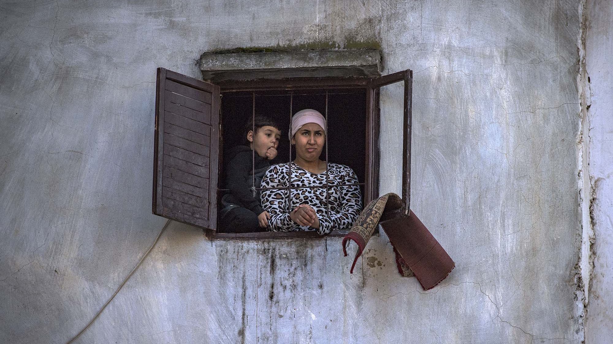 L'image montre deux personnes regardant à travers une fenêtre. Il y a une femme, vêtue d'un haut à motifs, qui semble attentive. À côté d'elle, un enfant, visiblement curieux, se penche légèrement. L'environnement est simple, avec des murs en crépi et une fenêtre aux volets ouverts. L'atmosphère semble réfléchie et calme.