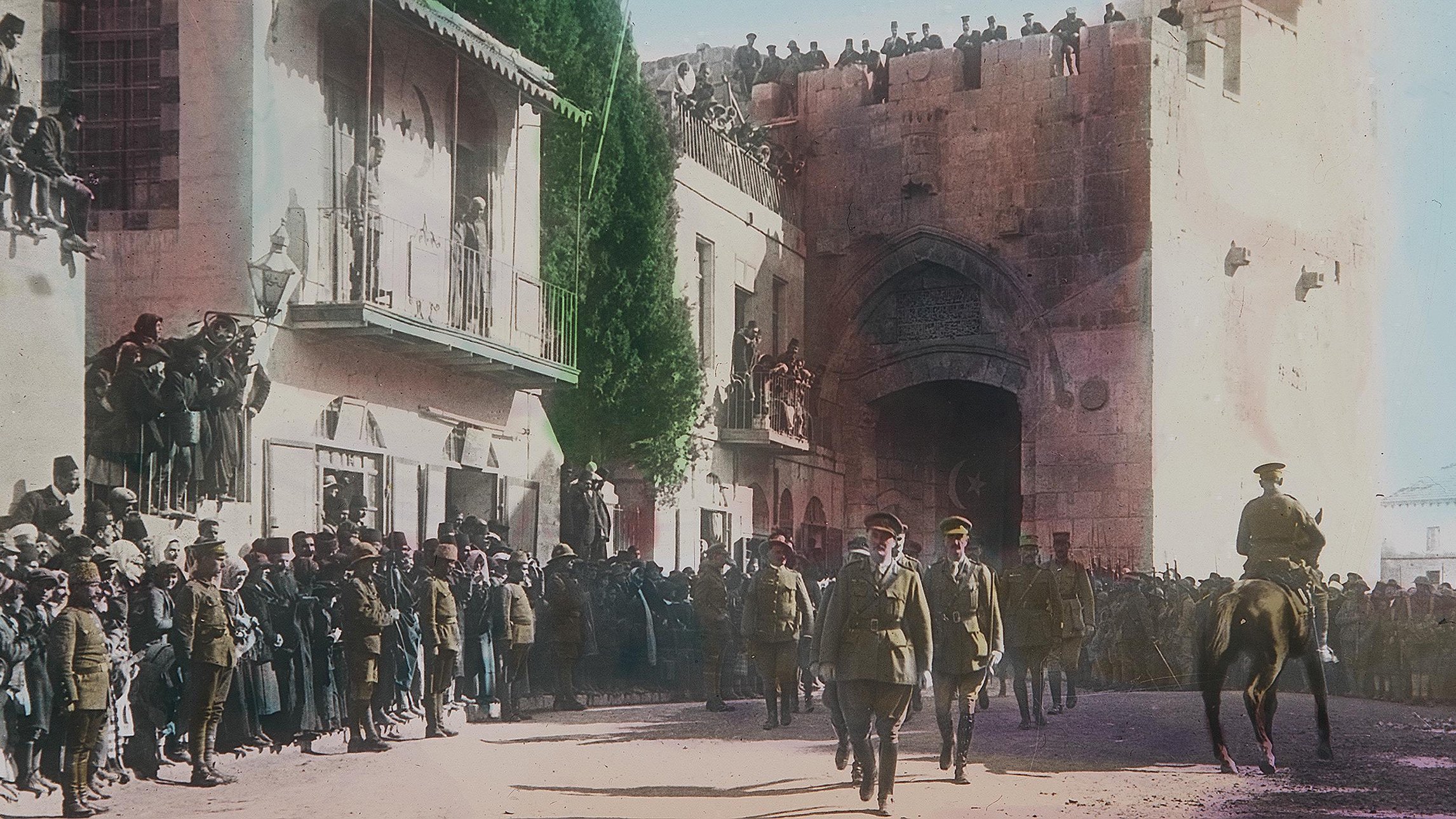 Cette image, en couleur, montre une scène historique se déroulant probablement lors d'un événement important à Jérusalem. Au premier plan, des soldats en uniforme, dont certains à cheval, avancent avec une certaine autorité. En arrière-plan, il y a une porte majestueuse sur un mur ancien, entourée de personnes qui regardent la procession depuis des balcons et des hauteurs. La foule est dense, avec des hommes, des femmes et des enfants, tous attentifs à ce qui se passe. Les bâtiments environnants sont typiques de l'architecture de la région, et l'atmosphère semble être à la fois festive et solennelle.