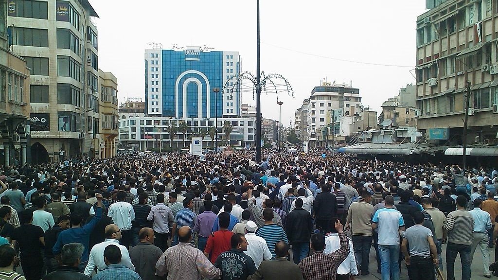 L'image montre une grande foule de personnes rassemblées dans une zone urbaine. On peut voir des bâtiments modernes en arrière-plan, ainsi qu'un aménagement urbain typique avec des lampadaires. La multitude semble engagée dans une manifestation ou un rassemblement, avec des gens de différentes orientations et tenues. L'ambiance générale évoque un moment de solidarité ou de protestation.
