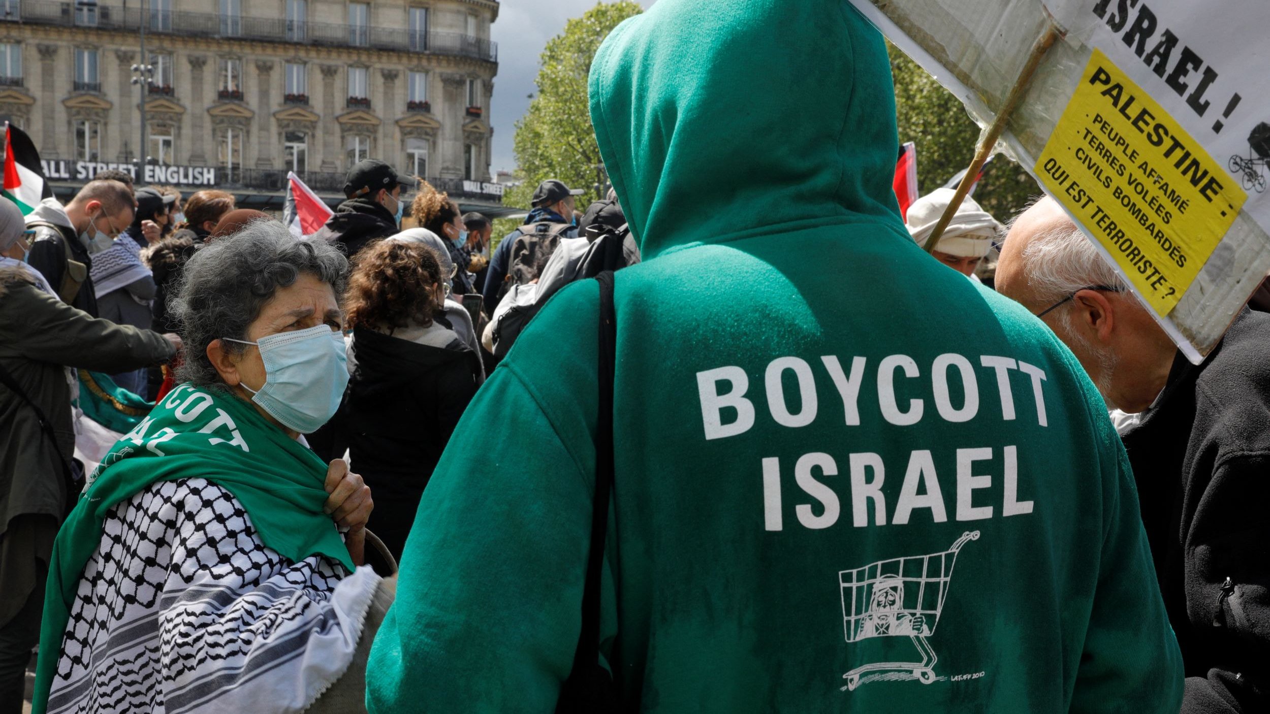 L'image montre une manifestation où des personnes se rassemblent pour exprimer leur soutien à la Palestine. Au premier plan, on voit une femme portant un foulard et un masque, conversant avec une autre personne. Celle-ci porte un sweatshirt vert sur lequel est inscrit "BOYCOTT ISRAEL". En arrière-plan, on distingue plusieurs personnes et des drapeaux, suggérant une mobilisation collective autour de cette cause. L'ambiance semble dynamique et engagée.
