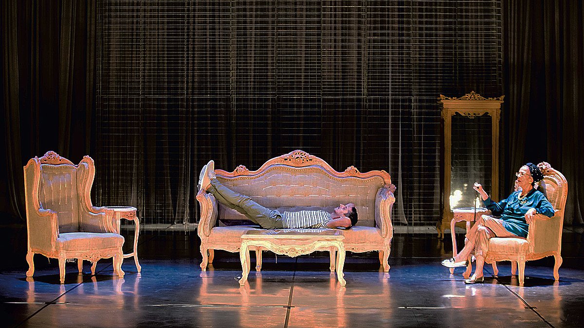 L'image montre une scène de théâtre avec un décor composé de plusieurs meubles anciens. Au centre, un sofa sur lequel un homme est allongé, semble détendu. À droite, une femme est assise sur une chaise, elle semble engagée dans une conversation ou une réflexion. Le fond est constitué d'un rideau sombre, ce qui met en valeur les meubles éclairés. L'ambiance semble à la fois intime et dramatique, suggérant une interaction entre les deux personnages.