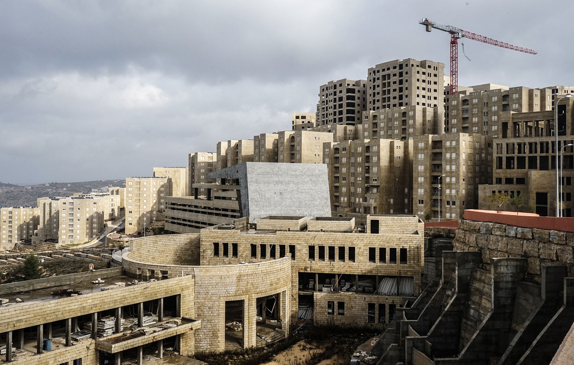 L'image montre un paysage urbain en construction, avec des bâtiments modernes de forme géométrique et des structures encore inachevées. On peut voir des façades en béton et des fenêtres non terminées sur plusieurs des bâtiments. En arrière-plan, une grue rouge indique que des travaux sont en cours. Le ciel est nuageux, créant une atmosphère un peu sombre. Au premier plan, des éléments de construction sont visibles, suggestifs d'un site en développement.