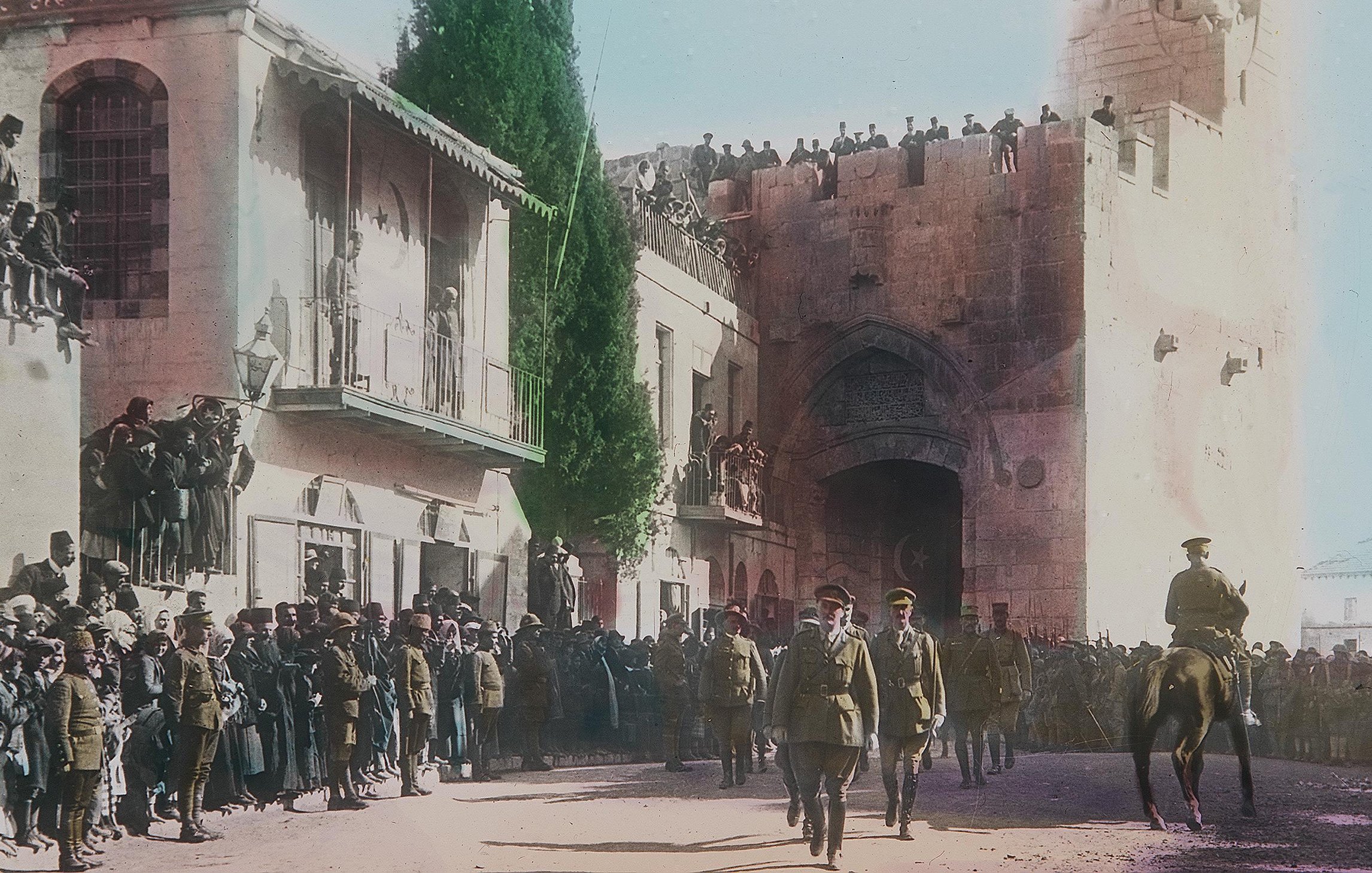 Cette image, en couleur, montre une scène historique se déroulant probablement lors d'un événement important à Jérusalem. Au premier plan, des soldats en uniforme, dont certains à cheval, avancent avec une certaine autorité. En arrière-plan, il y a une porte majestueuse sur un mur ancien, entourée de personnes qui regardent la procession depuis des balcons et des hauteurs. La foule est dense, avec des hommes, des femmes et des enfants, tous attentifs à ce qui se passe. Les bâtiments environnants sont typiques de l'architecture de la région, et l'atmosphère semble être à la fois festive et solennelle.