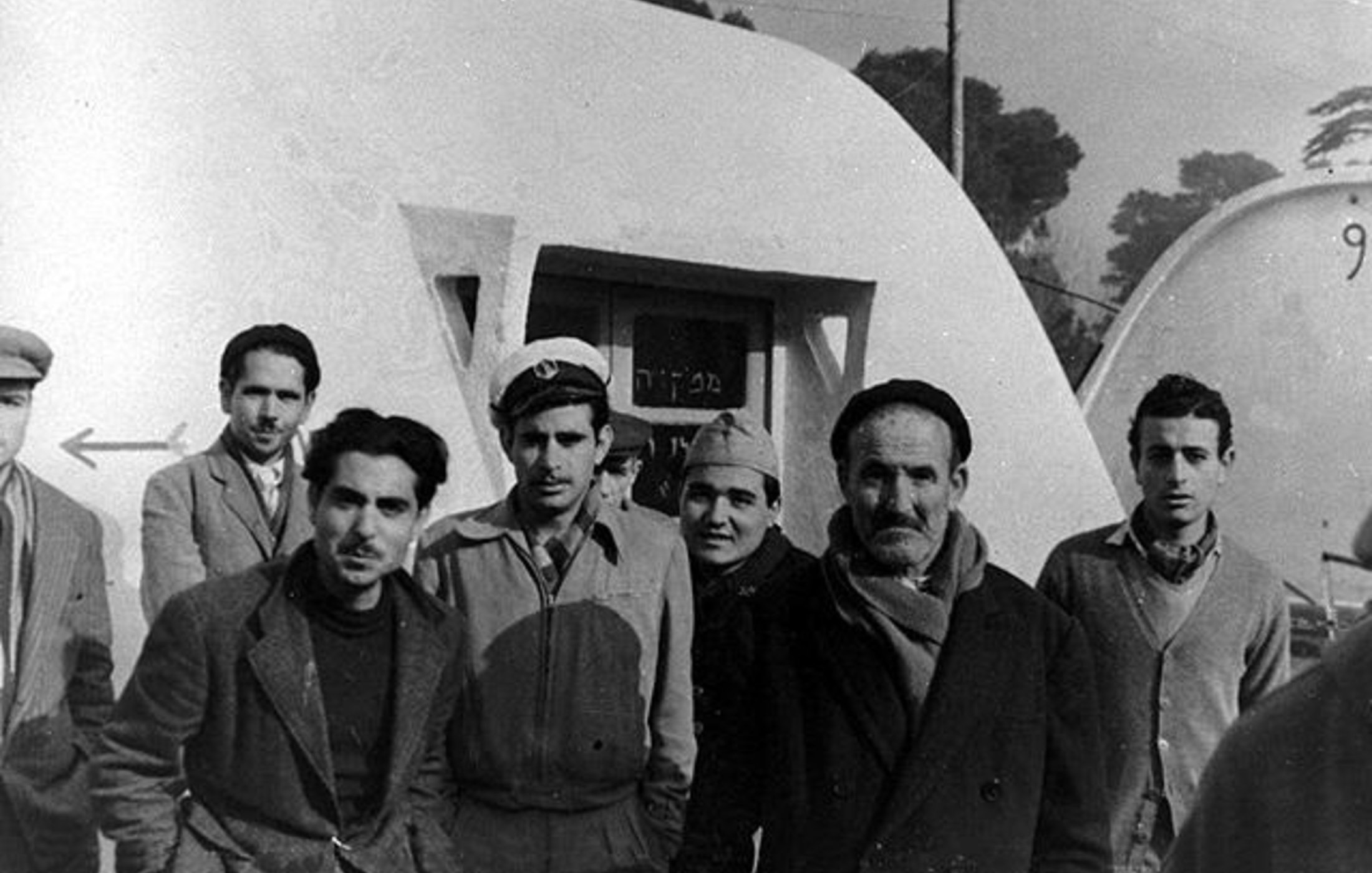 L'image montre un groupe de six hommes, debout près d'une structure blanche ressemblant à une maison ou une cabane. Ils sont habillés de manière variée, certains portant des chapeaux et des manteaux, ce qui suggère un temps frais. L'arrière-plan présente une autre structure similaire et des arbres flous. L'atmosphère semble décontractée, mais les expressions des hommes sont sérieuses.