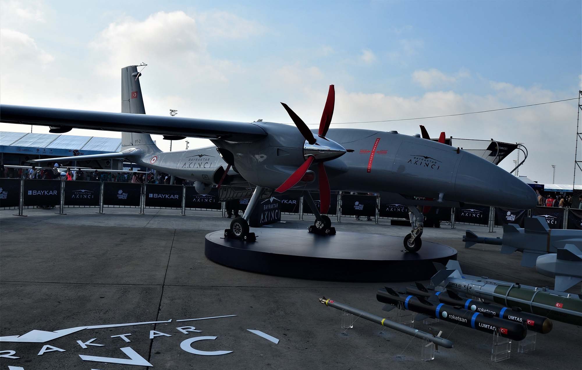 L'image montre un drone militaire, probablement de conception turque, exposé sur une plateforme de présentation. Le drone est gris avec des hélices rouges, et il possède des marquages distinctifs. À l'arrière-plan, on peut voir d'autres équipements, ainsi qu'une tente ou un pavillon. Au premier plan, il y a des missiles ou des armements disposés à côté du drone, indiquant qu'il s'agit d'une démonstration ou d'une exposition de matériel militaire. L'environnement semble être un salon aéronautique ou un événement similaire.