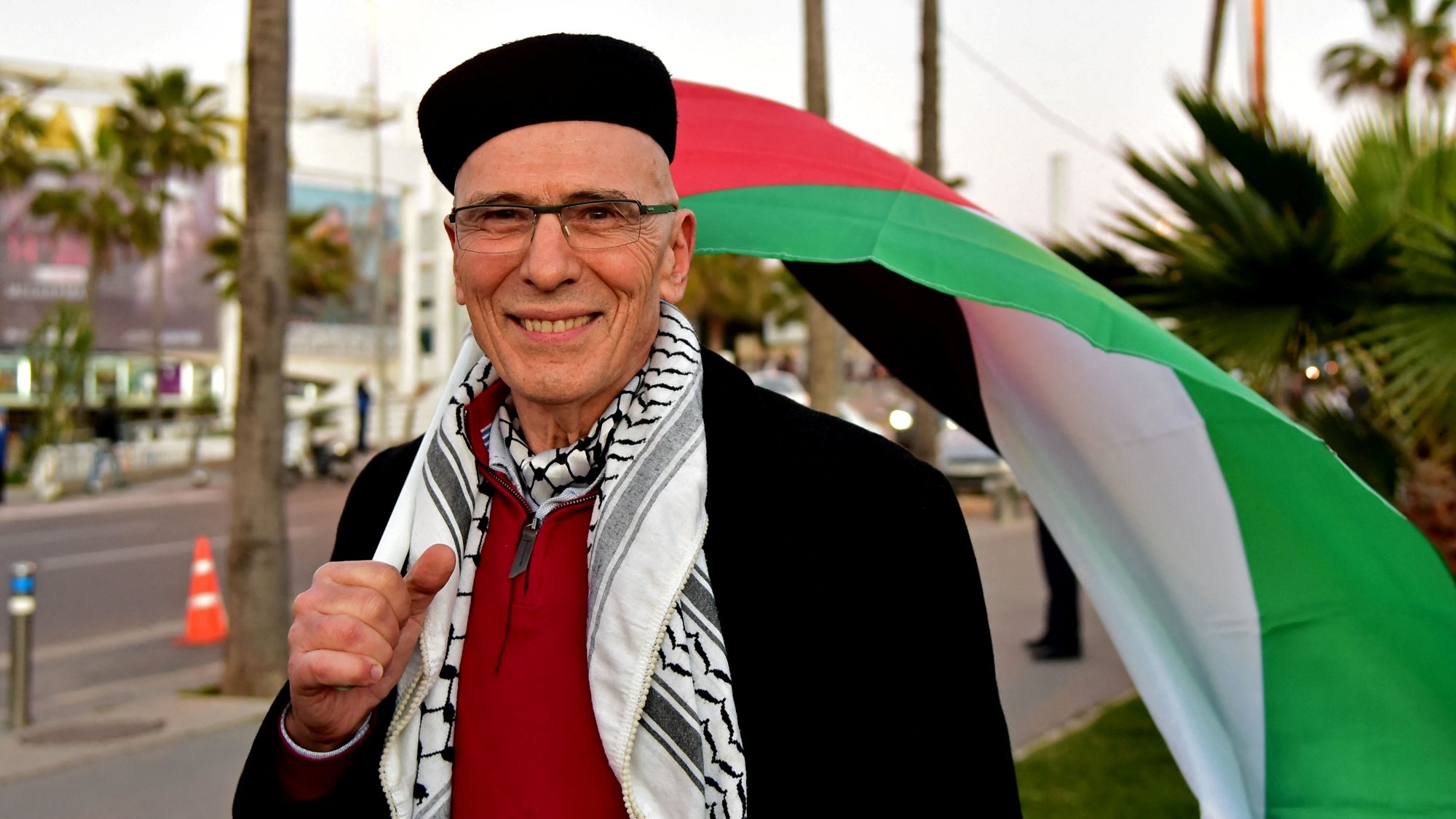 Un homme souriant porte un keffieh, derrière lui un drapeau palestinien sur fond urbain.
