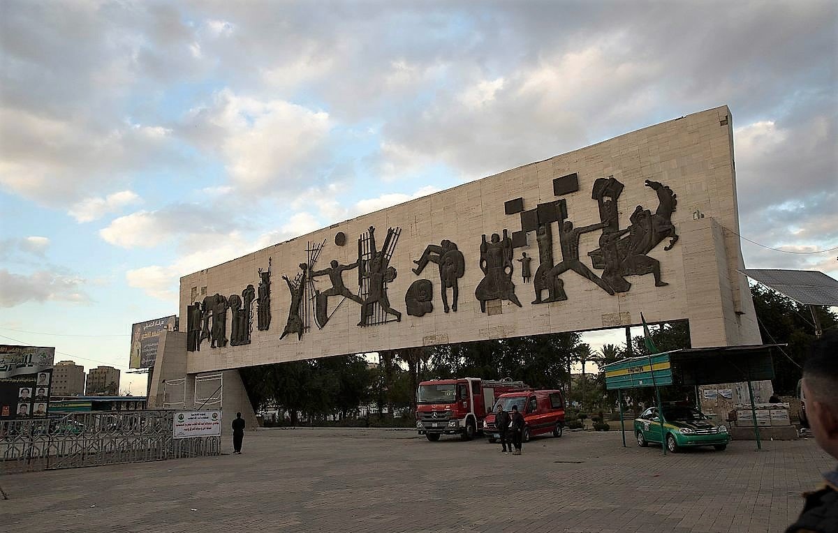 L'image montre une grande structure en béton surmontée de silhouettes découpées dans le matériau, représentant des figures humaines et des scènes de la vie quotidienne. Cette œuvre d'art moderne semble se dresser dans un espace public, entourée de véhicules et de personnes. Le ciel est nuageux, suggérant une ambiance calme et une lumière douce. En arrière-plan, on peut voir des bâtiments et des éléments d'urbanisme typiques d'une ville. L'ensemble évoque un mélange de culture et d'art, tout en étant intégré dans la vie citadine.