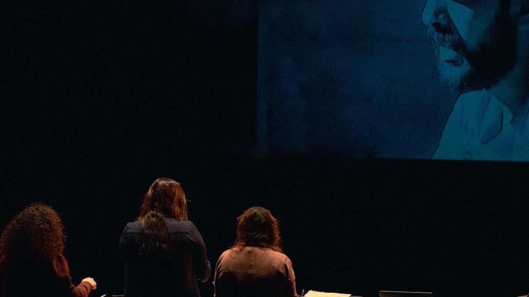 L'image montre un groupe de personnes de dos, se tenant devant une projection qui affiche une image en noir et blanc d'un homme portant un chapeau. Les personnes semblent engagées dans une activité, peut-être en lien avec la projection. Au premier plan, il y a un bureau avec un ordinateur et d'autres objets. L'éclairage crée une atmosphère intrigante autour de la scène.
