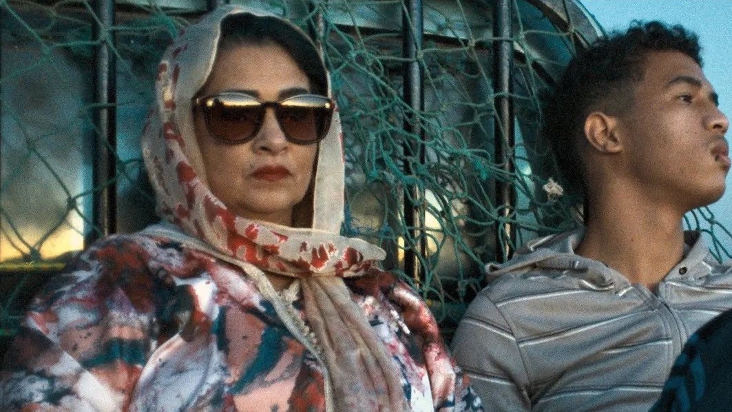 L'image montre une femme et un jeune homme assis ensemble dans un environnement désertique. La femme porte un foulard et des lunettes de soleil, affichant une expression attentive. Elle est vêtue d'une veste aux motifs colorés. À côté d'elle, le jeune homme porte un sweat à capuche et semble pensif, regardant au loin. En arrière-plan, on aperçoit des éléments qui évoquent un paysage aride. L'éclairage donne une atmosphère calme et contemplative à la scène.