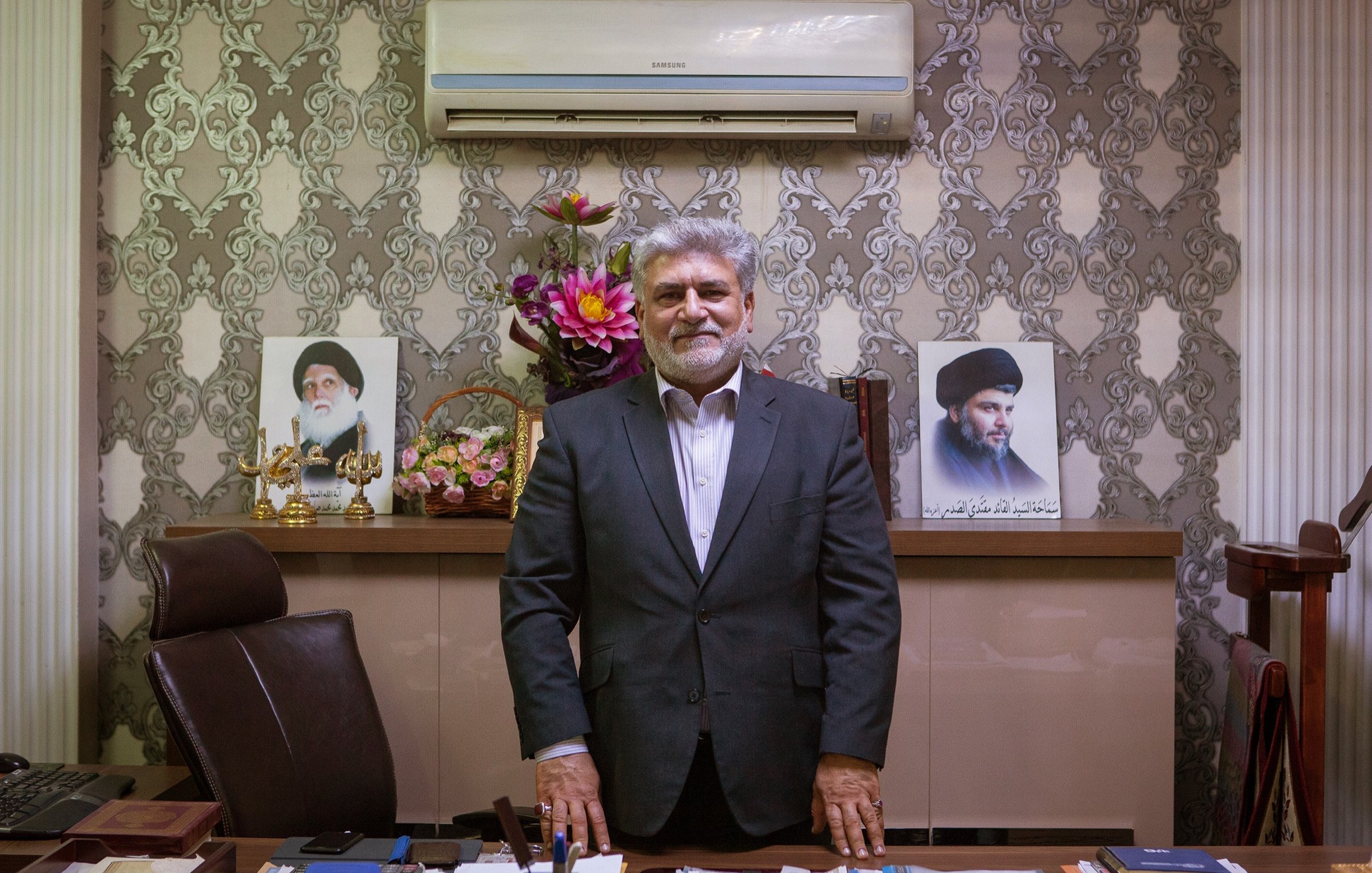 L'image montre un homme debout devant un bureau bien aménagé. Il porte un costume sombre et un sourire. Derrière lui, on peut voir des portraits de deux figures, ainsi qu'un arrangement floral. Le décor est élégant avec des murs ornés de motifs. L'ensemble dégage une atmosphère de professionnalisme et d'autorité.