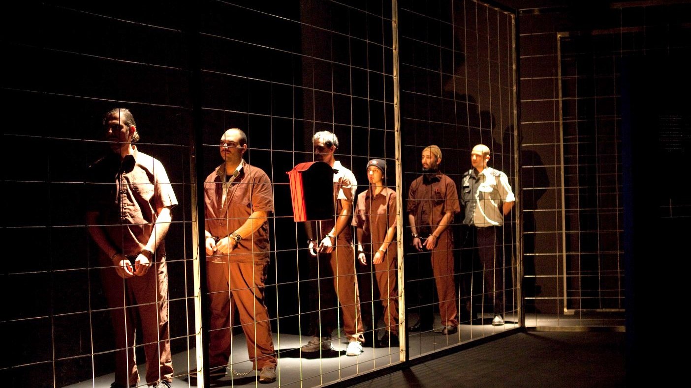 L'image montre un groupe d'hommes disposés en ligne, chacun portant des vêtements de style uniforme. Ils se tiennent derrière un grillage, ce qui crée une impression de séparation ou de détention. L'éclairage est sombre, mettant en valeur les figures des hommes, qui semblent concentrés ou pensifs. L'ambiance générale évoque des thèmes de confinement et d'isolement.