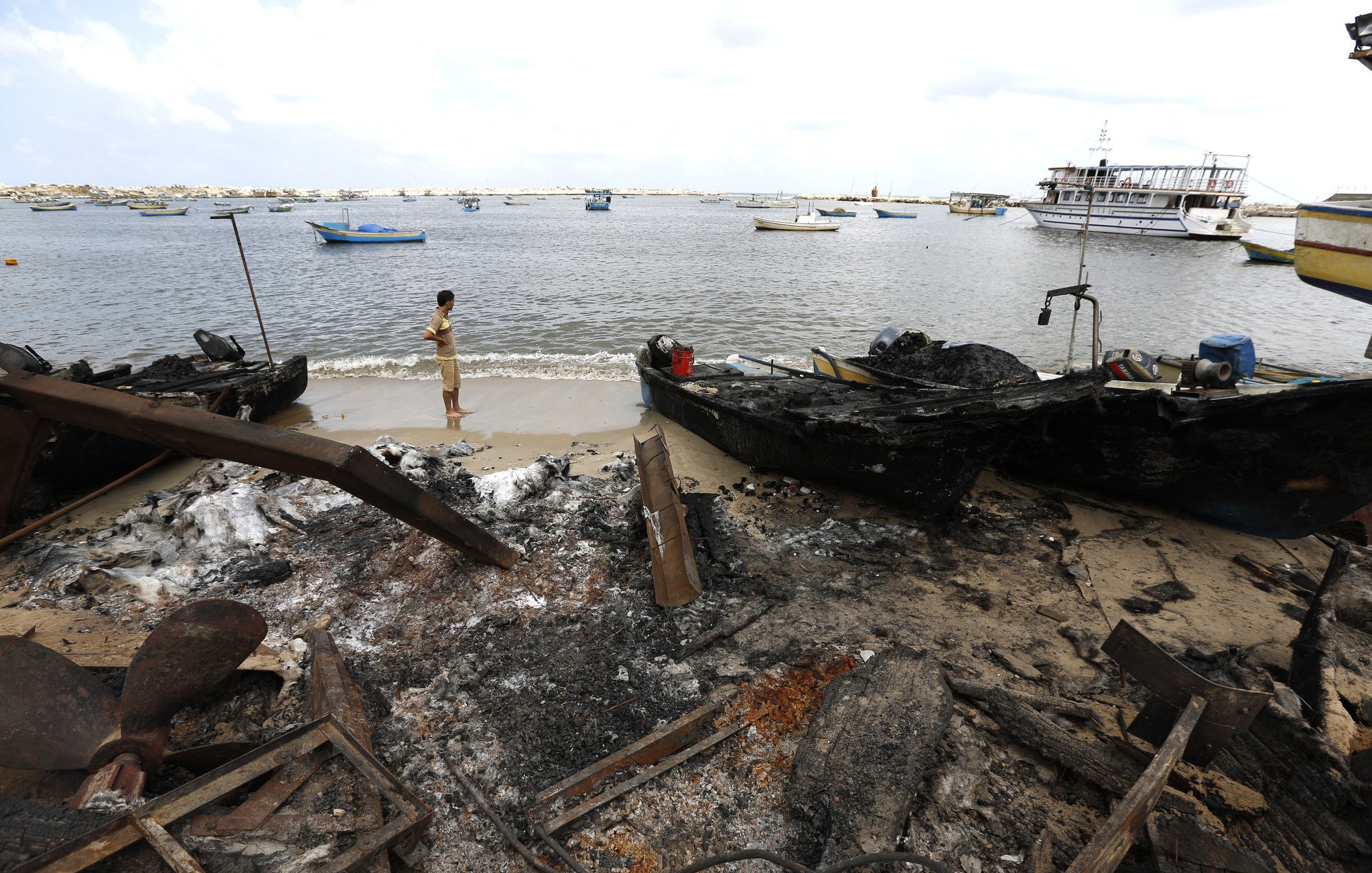 L'image montre une scène côtière après un incendie. On peut y voir des bateaux endommagés et carbonisés sur le rivage. Un homme se tient près de l'eau, regardant vers le horizon. La mer semble calme, avec plusieurs petites embarcations au loin. Le ciel est nuageux, et le paysage est marqué par les conséquences de l'incendie, avec des débris et des cendres sur le sable.