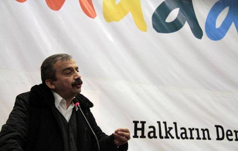 L'image montre un homme parlant lors d'un événement ou d'une conférence. Il est vêtu d'un manteau sombre et d'une chemise blanche. Le fond affiche une grande bannière avec des mots colorés, dont "Halkların Derneği", qui signifie "L'Association des Peuples" en turc. L'homme semble engagé dans son discours, utilisant des gestes pour illustrer ses propos.