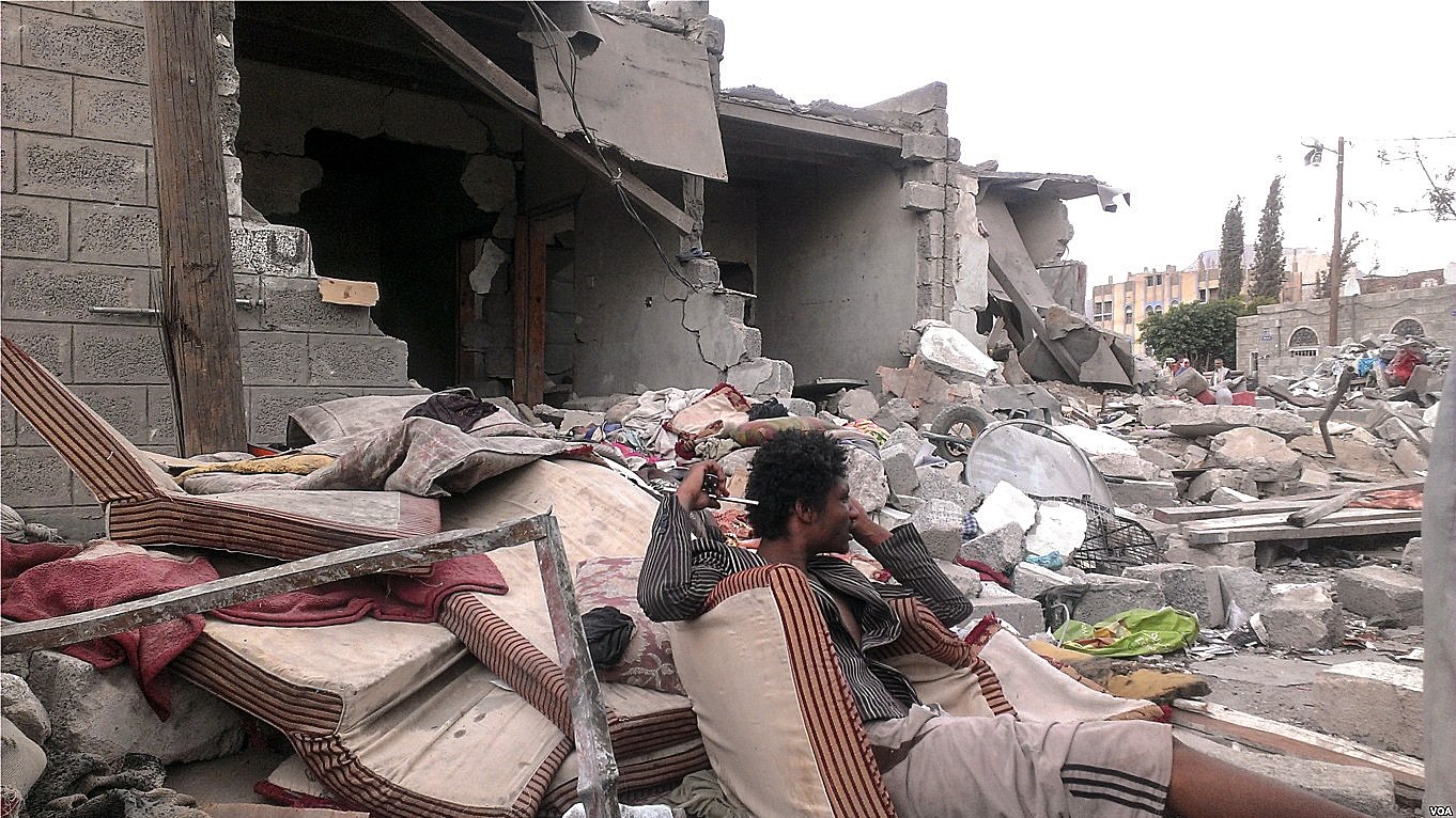L'image montre une scène de destruction, probablement après un conflit ou une catastrophe naturelle. Au premier plan, un homme assis sur des matelas usés semble perdu dans ses pensées, entouré de débris. À l'arrière-plan, des bâtiments partiellement effondrés et des gravats jonchent le sol, témoignant des conséquences dévastatrices de l'événement. L'atmosphère est sombre et tragique, avec une sensation de désolation.