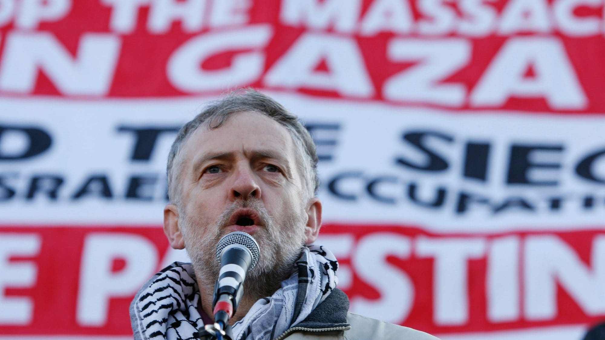 L'image montre un homme prenant la parole devant une grande banderole. Il porte un foulard traditionnel autour du cou et semble s'adresser à une foule. La banderole en arrière-plan contient des messages concernant la situation à Gaza, appelant à mettre fin aux massacres et à la occupation actuelle. L'expression de l'homme indique qu'il est engagé et passionné par le sujet qu'il aborde.